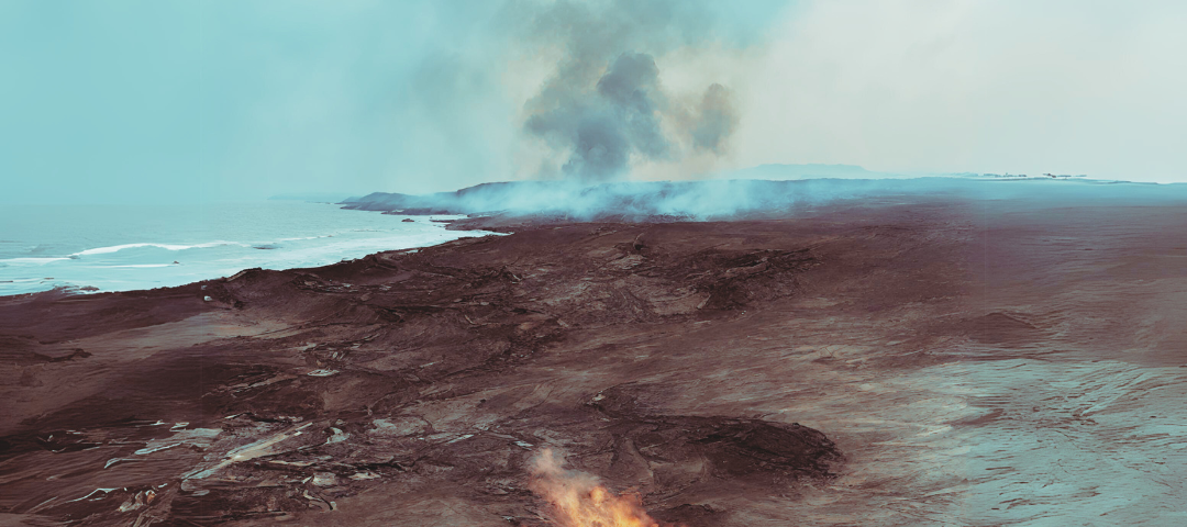 A small fire smoulders on a piece of arid land, with the sea in the distance, and smoke and ash floating in the air.