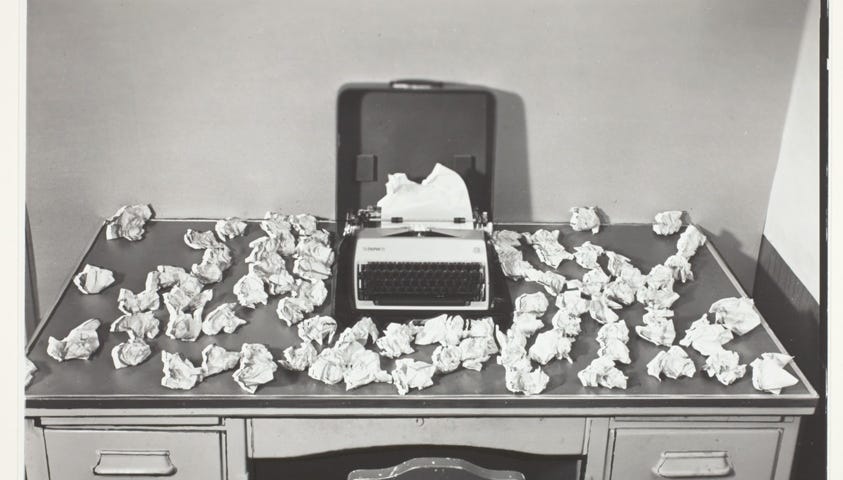 blac and white photo of an old desk littered with crumpled sheets of paper surrounding a typewriter. (words for writers)