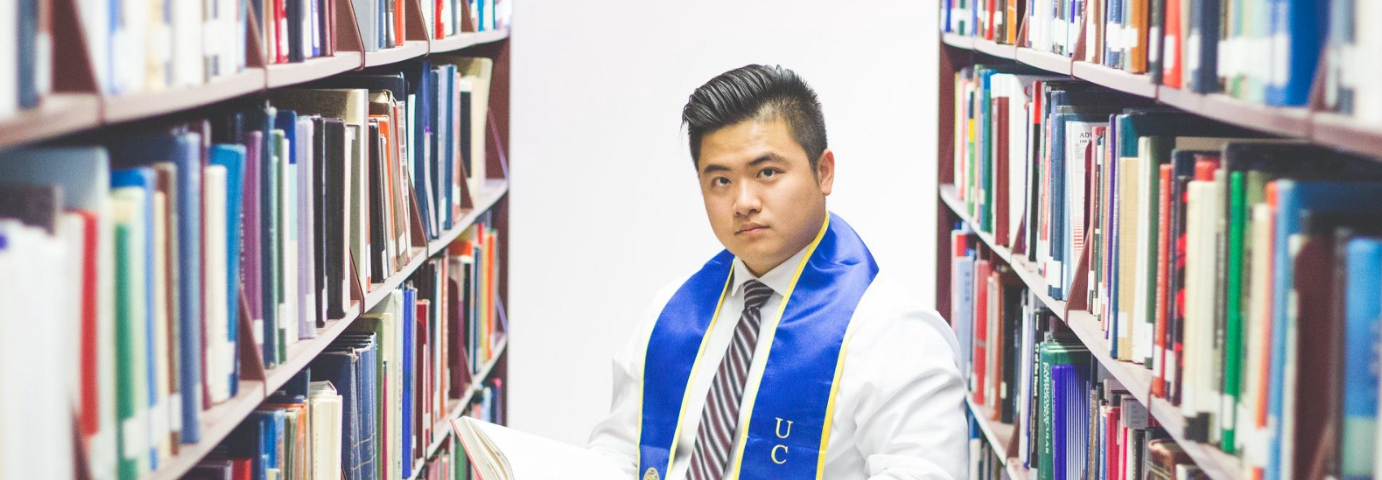 A UC Irvine student looks at books in the library, wearing their graduation regalia.