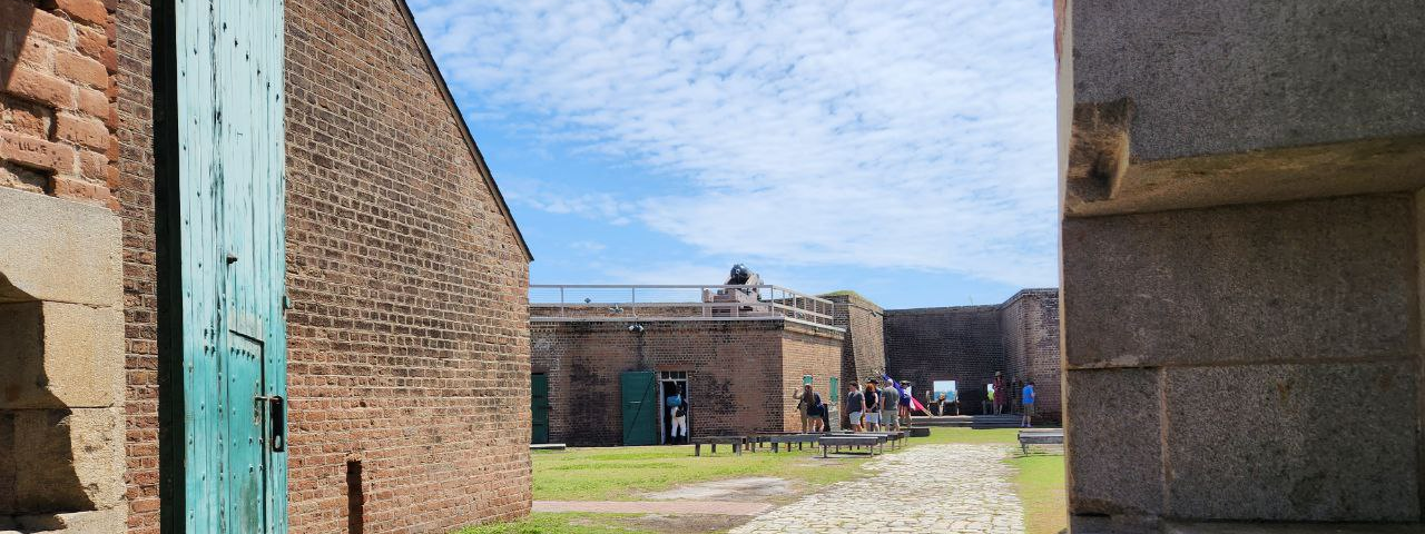Interior of a historic brick fort with cobblestone pathway, a green wooden door, and an upper wraparound level with cannons in the distance