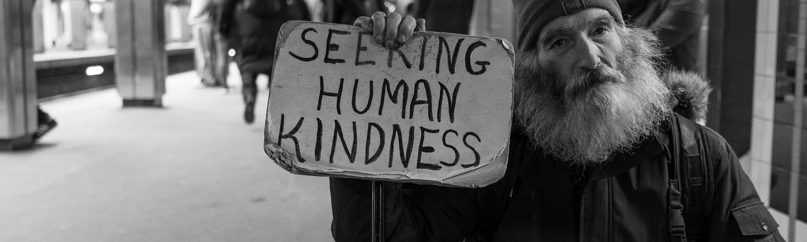 A bearded man holding a sign that reads “Seeking human Kindness”.