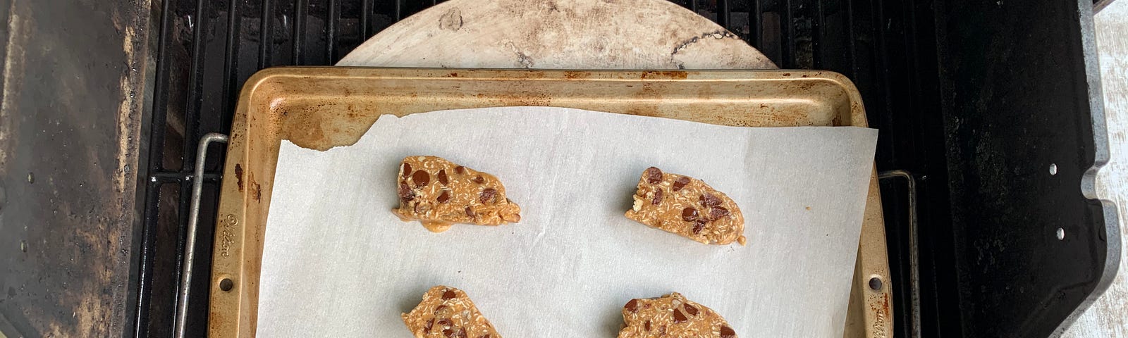 Slices of chocolate chip cookie dough set on a parchment paper lined cookie sheet atop a pizza stone on top of a BBQ grill.