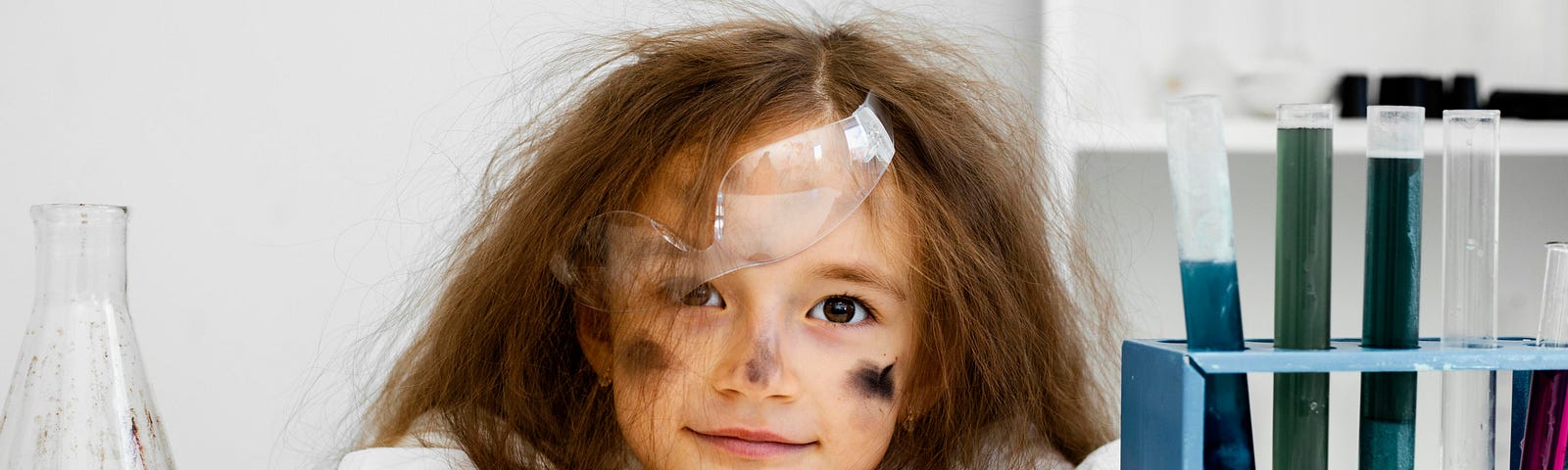 Little girl with smoke on her face, test tubes beside her and protective glasses halfway off her face