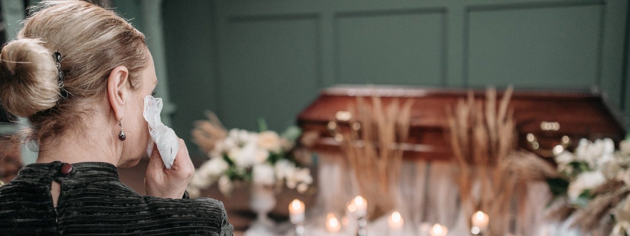 A blonde woman dabs her eyes with a tissue. She is sitting on a church bench near a closed casket surrounded by candles and flowers.