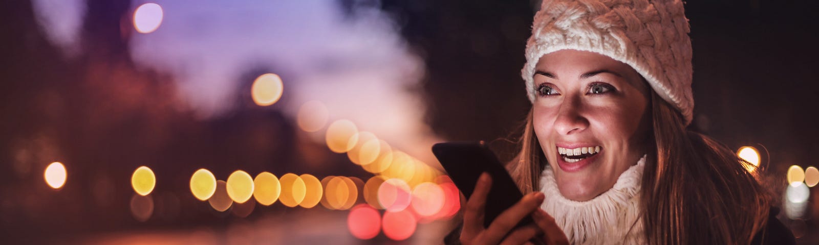 photo of a smiling, young, vibrant woman wearing a knitted woolen hat and chunky, warm textured sweater and warm coat holding her phone in front of her mouth and talking into it, against a backdrop of a blurred city streetscape at dusk.