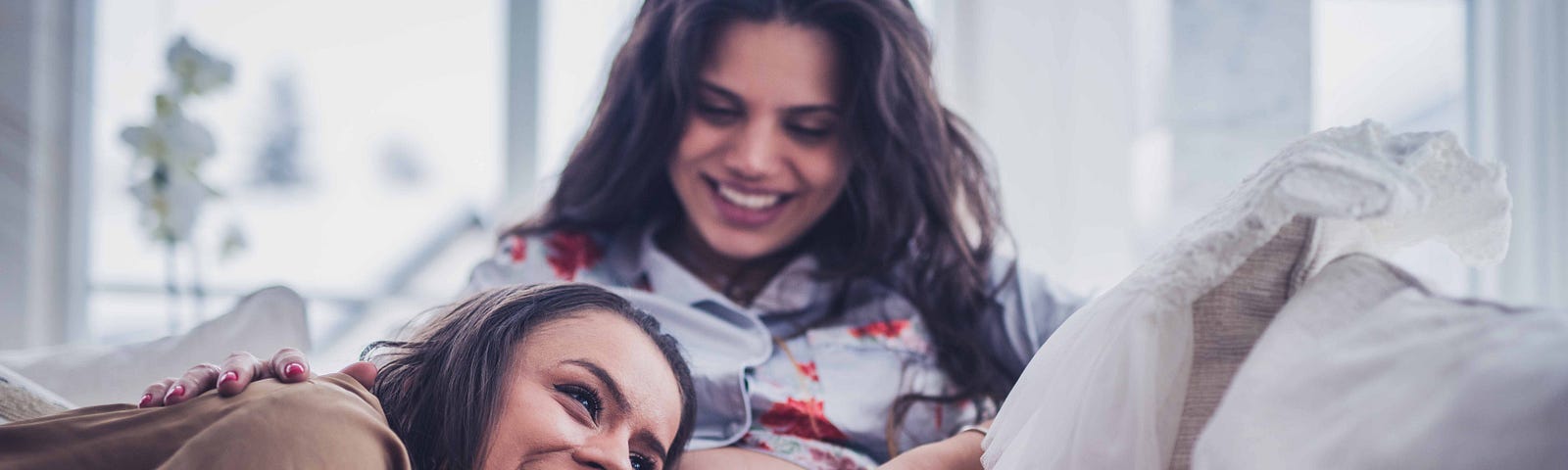 Pregnant woman and another woman sitting on a couch in a living room laughing together as one woman rests her head on the pre