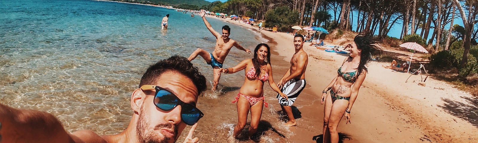 A smiling twenty-something guy on a sunny beach takes a selfie pointing at all his cool, glamorous and smiling friends in the background