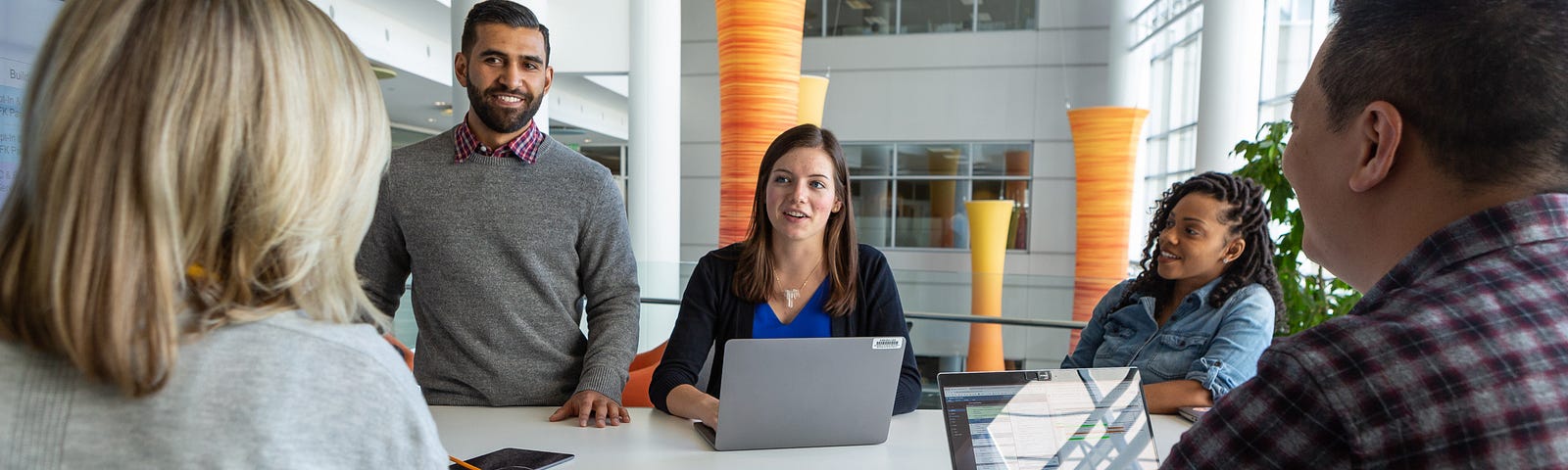 A group of five colleagues sit around a table in an office with open laptops, collaborating and talking