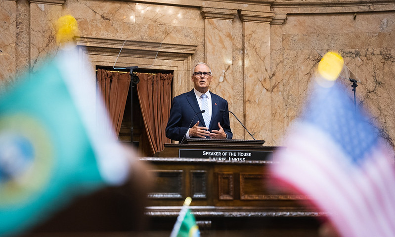 Photo of Jay Inslee in the House chambers speaking from the rostrum. Blurred images of the Washington state and U.S. flags frame the foreground.