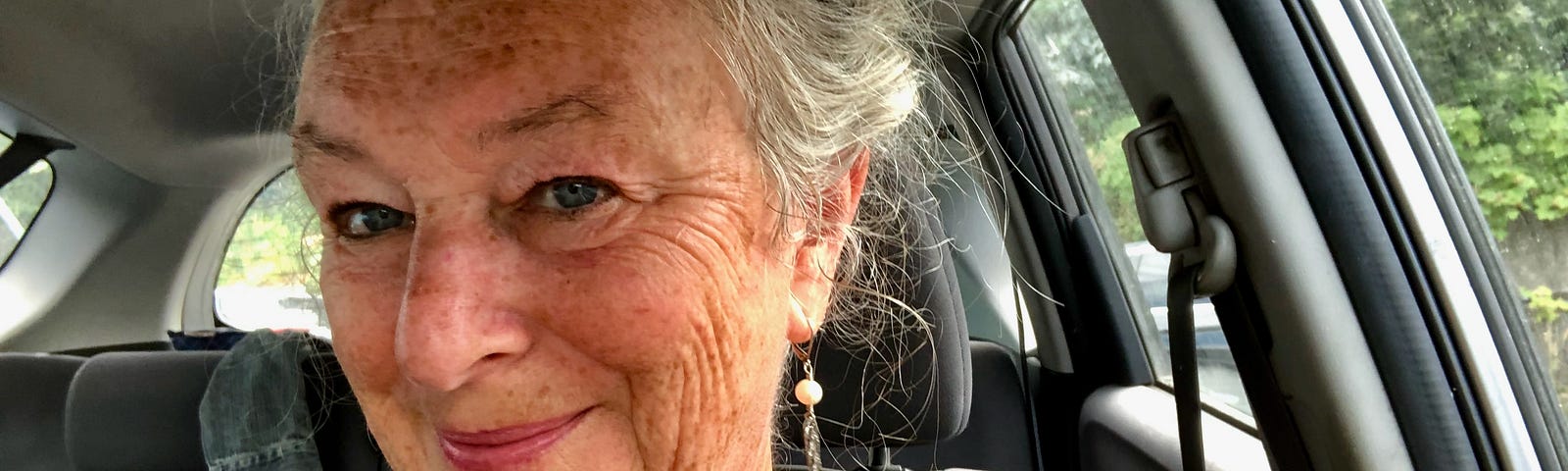 An older woman sits in a car. She is smiling at the camera. She has grey hair and many wrinkles on her face and neck