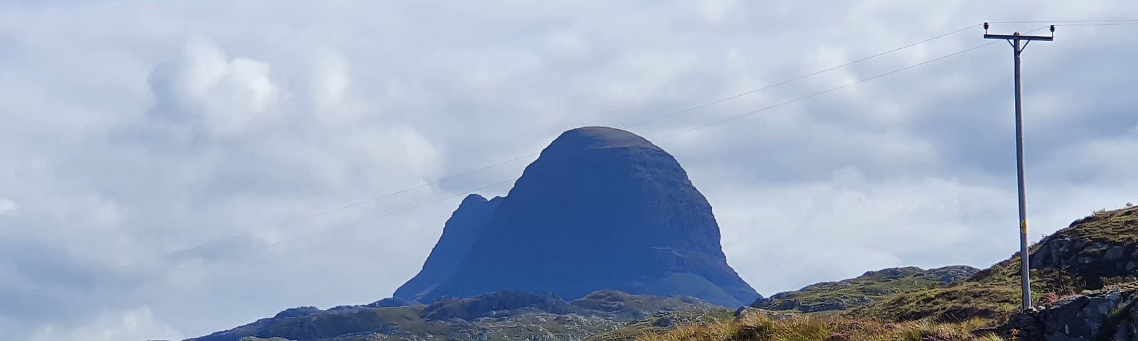 This shows the distinct hump silhouetted outline of a mountain called Suilven black against the summer sky and towering above the surrounding area which is covered with heather.