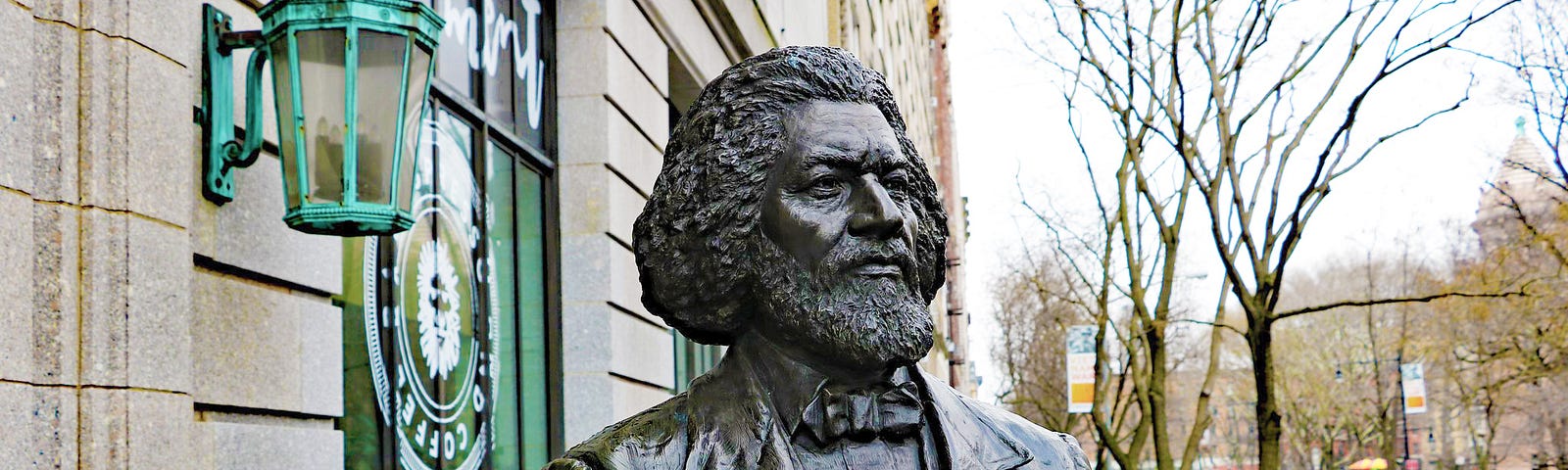 Bronze statue of a man from the nineteenth century. He’s looking forward, and stands next to a decorative copper lamp.