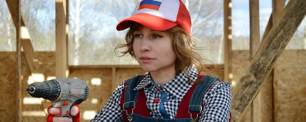 An attractive woman sitting on a construction site. She is wearing a blue and white check shirt, denim dungarees, red gloves, and a red and white baseball cap. She is holding a drill.