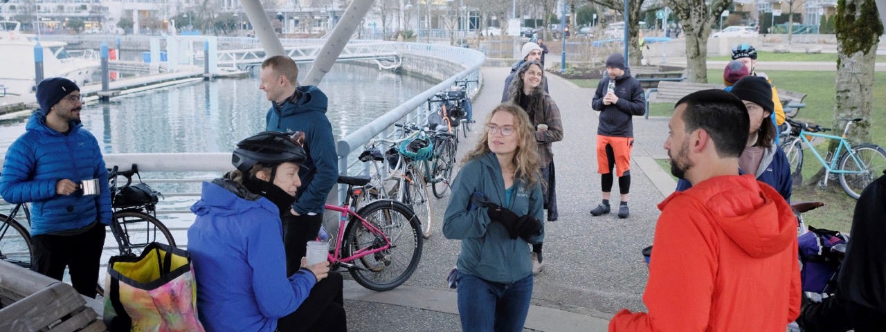 A group of cyclists drinking coffee and chatting at a waterfront park in downtown Vancouver. Behind the cyclists are five bicycles, leaning against a railing.