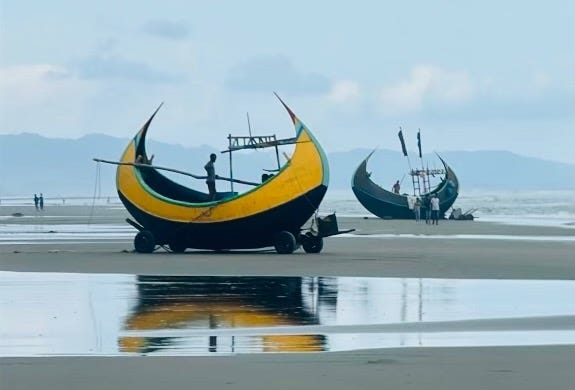 Two crescent shaped moon boats sit on a flat sand beach in Bangladesh. One is bright yellow and the one further back is blue.