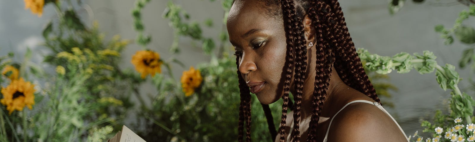 A Black woman with box braids reads a book, behind her are yellow flowers.