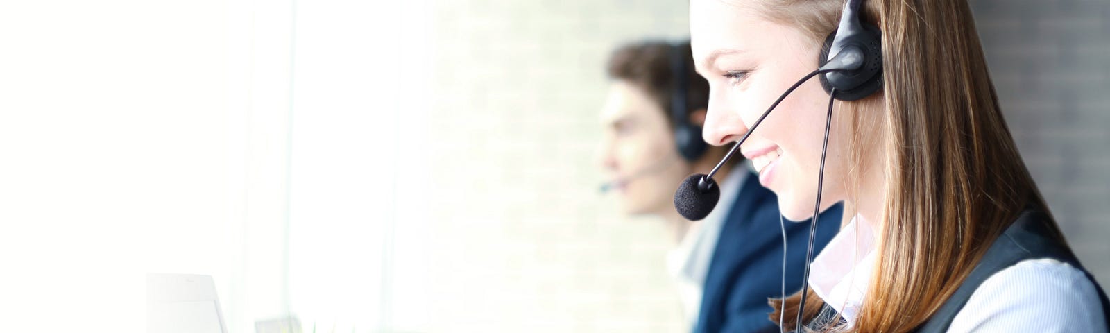 A woman with a headset smiles at a computer. A blurry man also wearing a headset can be seen sitting behind her.