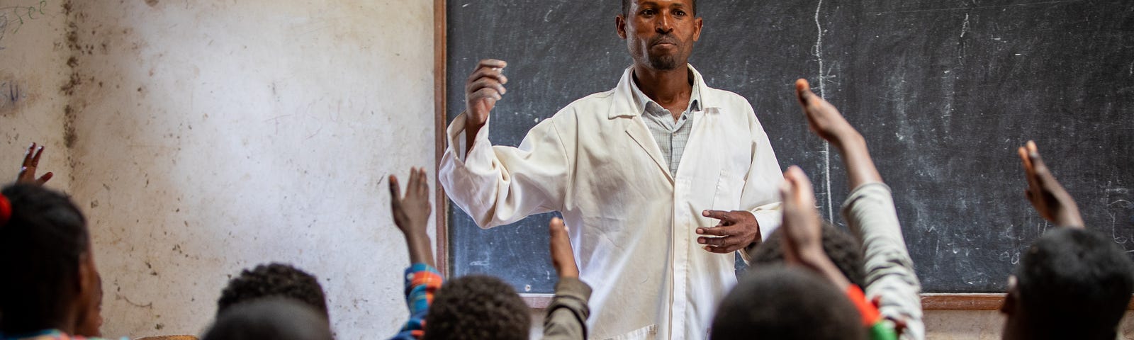 A man wearing a white lab coat stands in front of a blackboard in a room full of children sitting at desks and raising their hands.