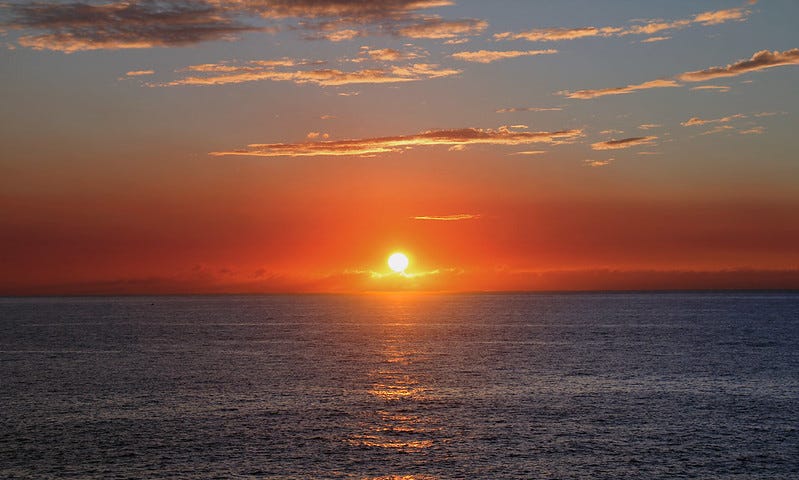 A view over dark blue water of the sun near the horizon, appearing red, with orange-lit clouds above.