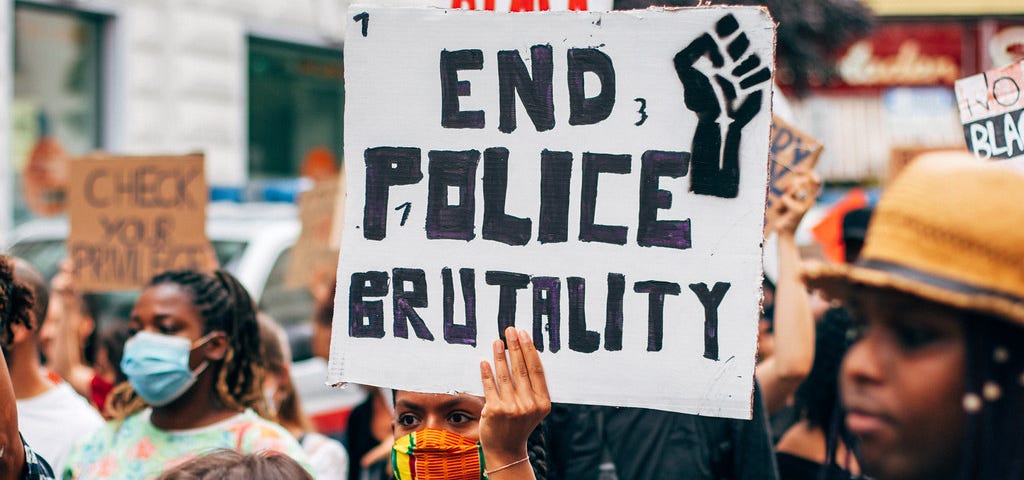 An image of a crowd of protesters. In the center, a woman with brown skin and a colorful face masks holds up a white sign with “End Police Brutality” in black writing and a Black Power fist.