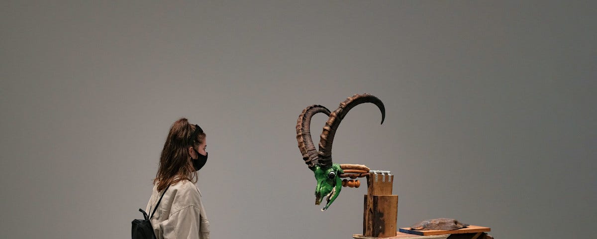 A woman wearing a small backpack and a facemask is standing opposite an art installation which seems to be a four-legged table made from broken chairs and other wooden junk, with a wooden block on top to form a neck, to which is attached a green-painted goat skull. The skull has large horns.