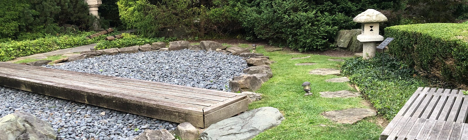 A circular rock garden with wooden planks across it, surrounded by shrubs and grass. Two wooden benches are on the right