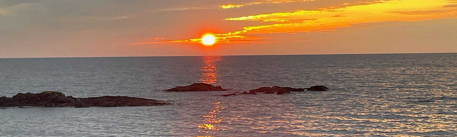 Orange sun in a yellow and orange sky is on the horizon, casting sun light on the lake and rocks below.