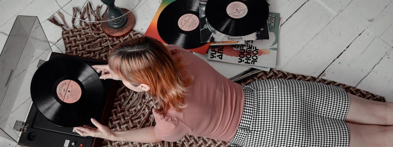 Woman lying on the floor, putting a record on her turntable, guitar and other records next to her.