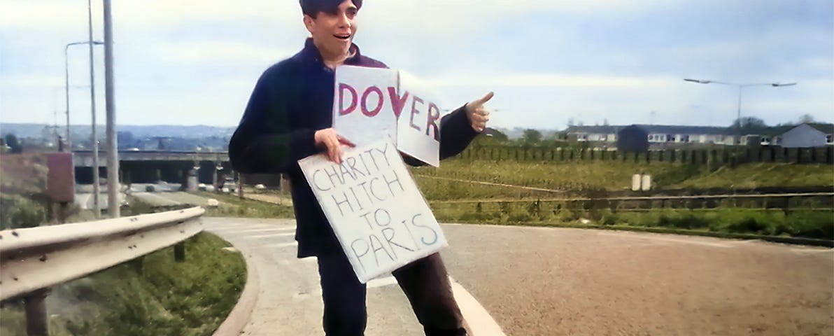A young man hitchhiking on a English motorway. He is holding two signs that say ‘Dover’ and ‘Charity Hitch to Paris.