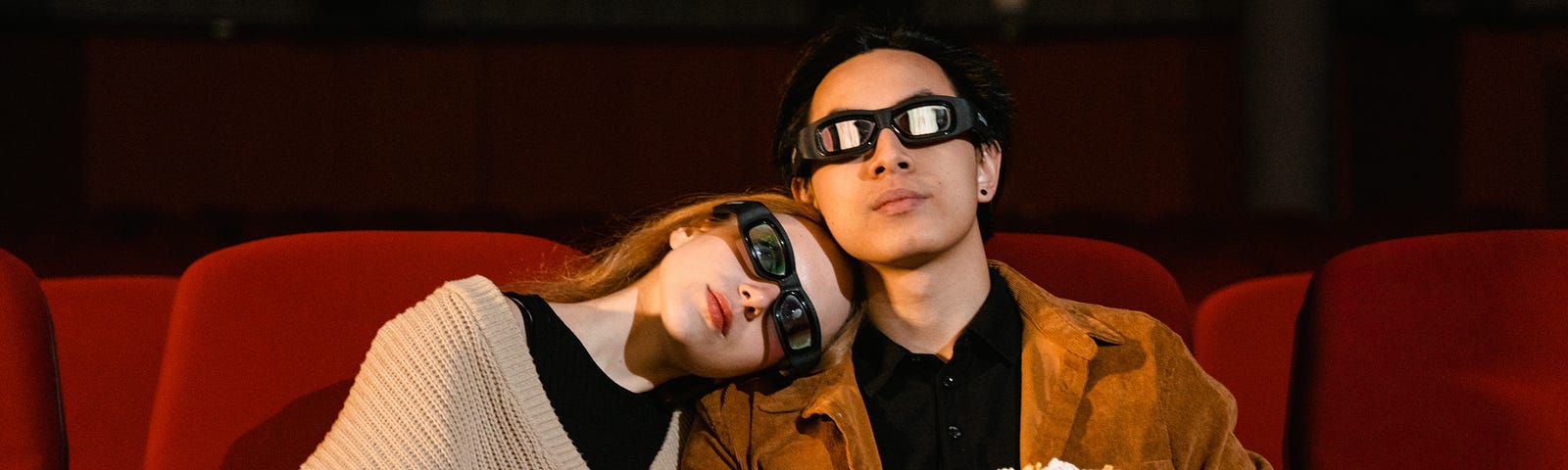 A couple watching a movie at the cinema, wearing 3D glasses and holding popcorn.