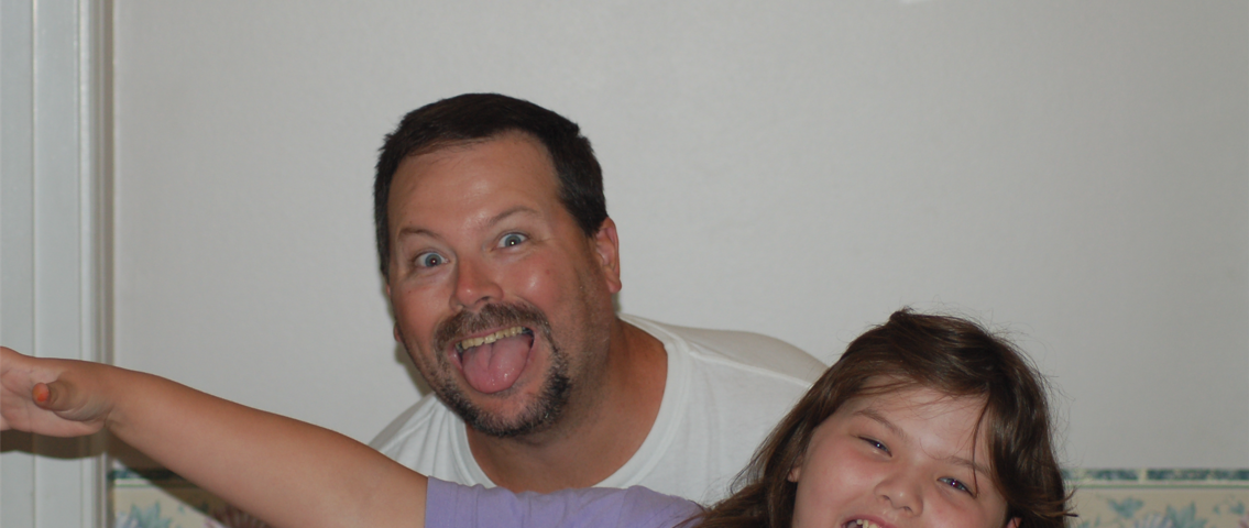 A picture of a middle-aged man and his young daughter, both brunet and tan with similar features. They’re posing for the camera and making goofy expressions. There’s dated wallpaper behind them with a seashell motif.