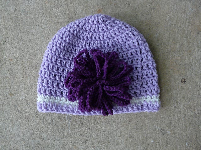 A light purple crochet hat with one strip of white toward the bottom and big, purple flower on the side