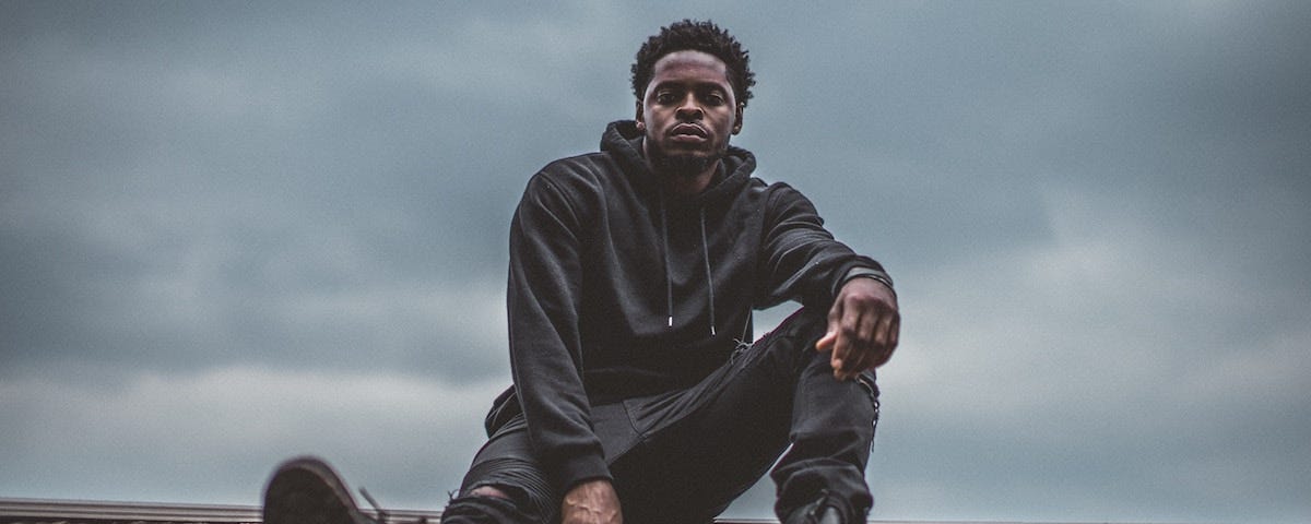 Black man wearing all black clothing sitting on top of a roof with cloudy grey skies behind him. A photo by Chase Fade.