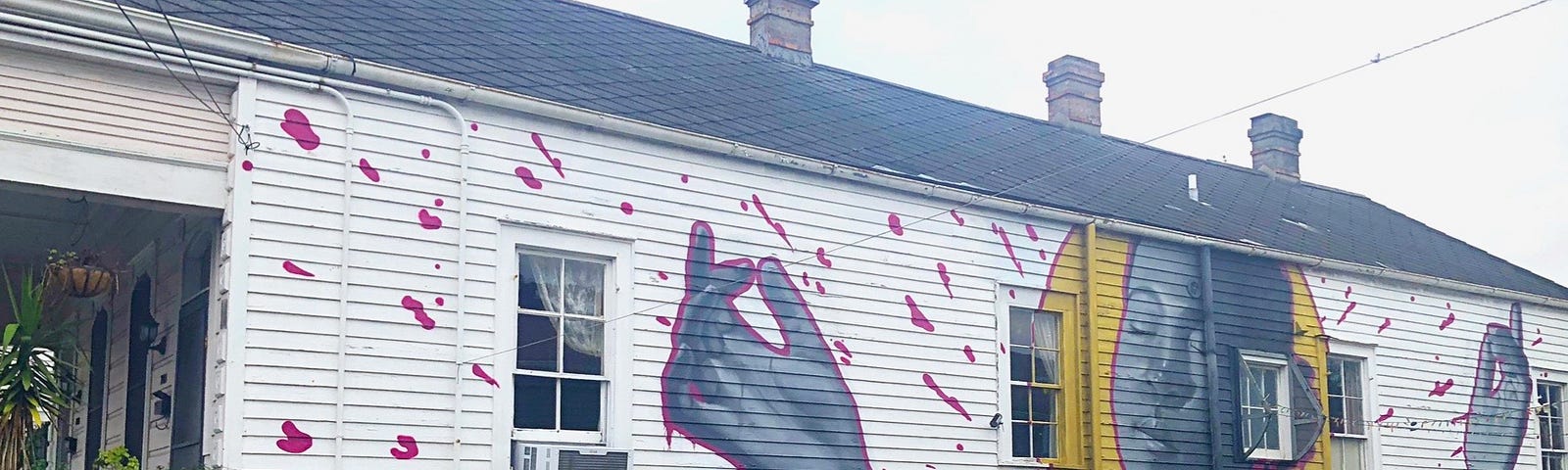 Long, white clapboard house with 3 chimneys, the side is covered by a painting of the upper body of a black woman — eyes closed, hand reaching up to pluck a miracle, a blue butterfly at her throat chakra, and a gold halo behind her head.