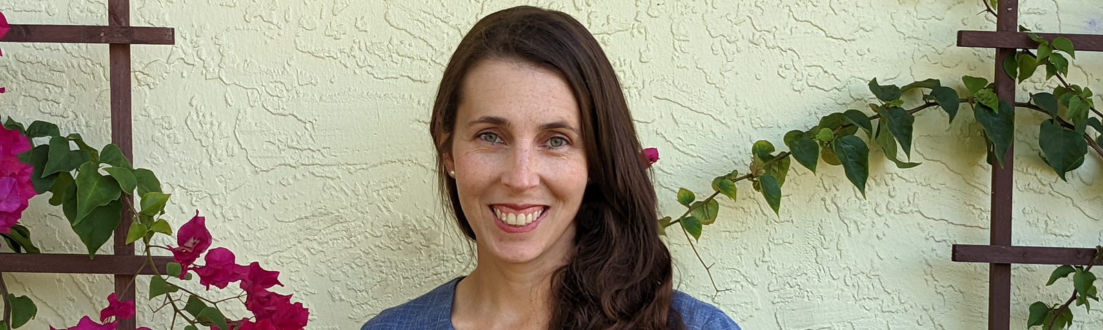 Wearing a blue v-neck blouse with her long, dark hair parted and draped over the front of her left shoulder, Marilys Galindo is pictured in front of a bright, stucco wall with flowering vines trained to provide a colorful but undistracting backdrop to her warm smile.