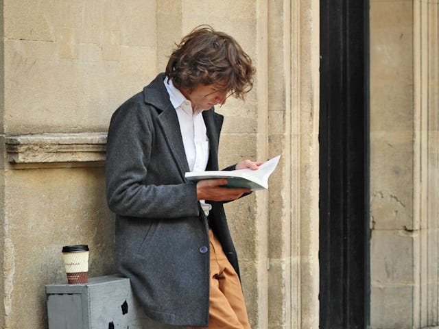 A man standing against a sone wall holding a book in both hands with a coffee at his side.