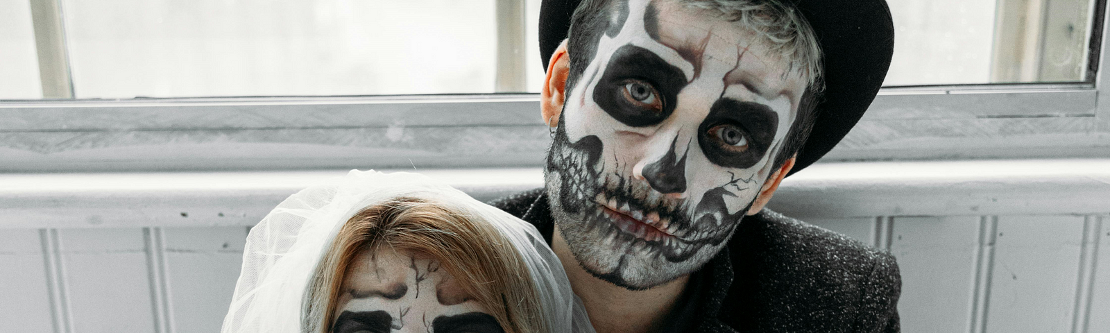 A man and woman both with painted skull faces and looking like a bride and groom. The man is sitting against a wall with a window behind. It is daylight. he is looking into the camera. The woman is sitting between his legs looking away. She doesn’t look very scared but the man does have his hands gently around her neck.