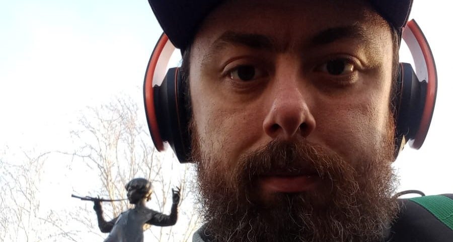 A bearded man with headphones on in front of a statue of a boy playing the flute