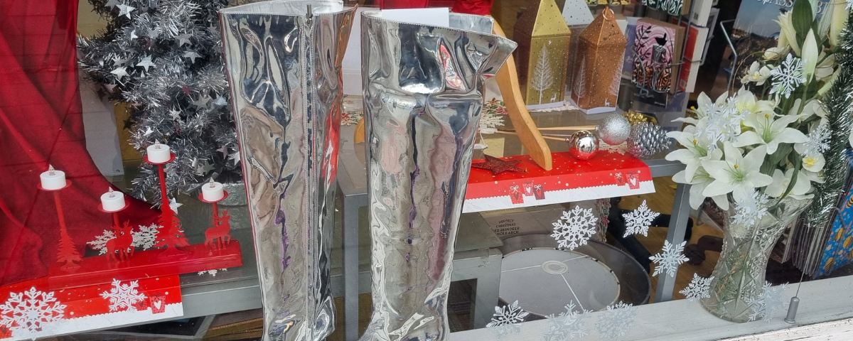 A photo of over-the-knee shiny silver high heeled boots in the window of a charity shop. The window has a sparkly festive look.