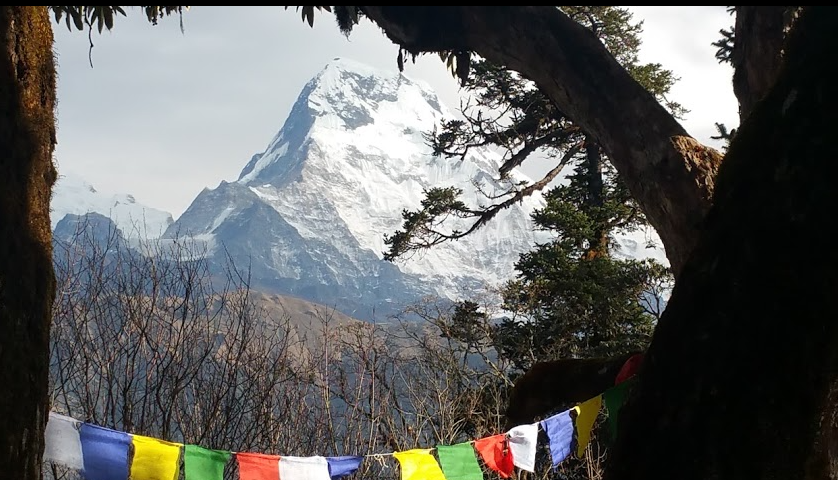 Looking out from a gap in the trees at a huge, snow capped mountain peak in the distance. In the foreground, Buddhist Prayer Flags are strewn between the trees. Coloured blue, yellow, green, red and white.