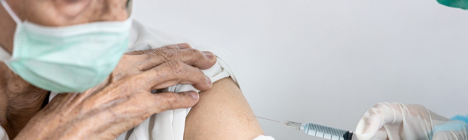 An elderly woman in a white top and mint disposable mask with her sleeve rolled up, getting an injection/vaccination from a doctor/nurse in full protective gear.