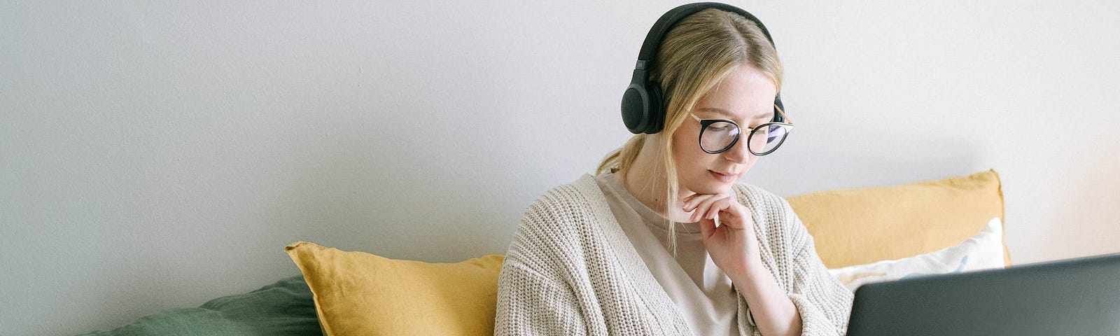 Photo of Woman wearing classes is Sitting on a Couch or Bed and Writing in her Notebook while her Laptop is open