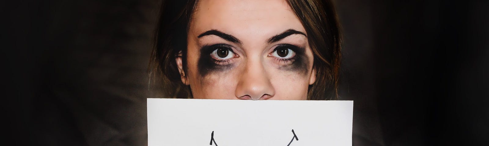 Woman with smeared mascara holds a drawing of a smile in front of her face
