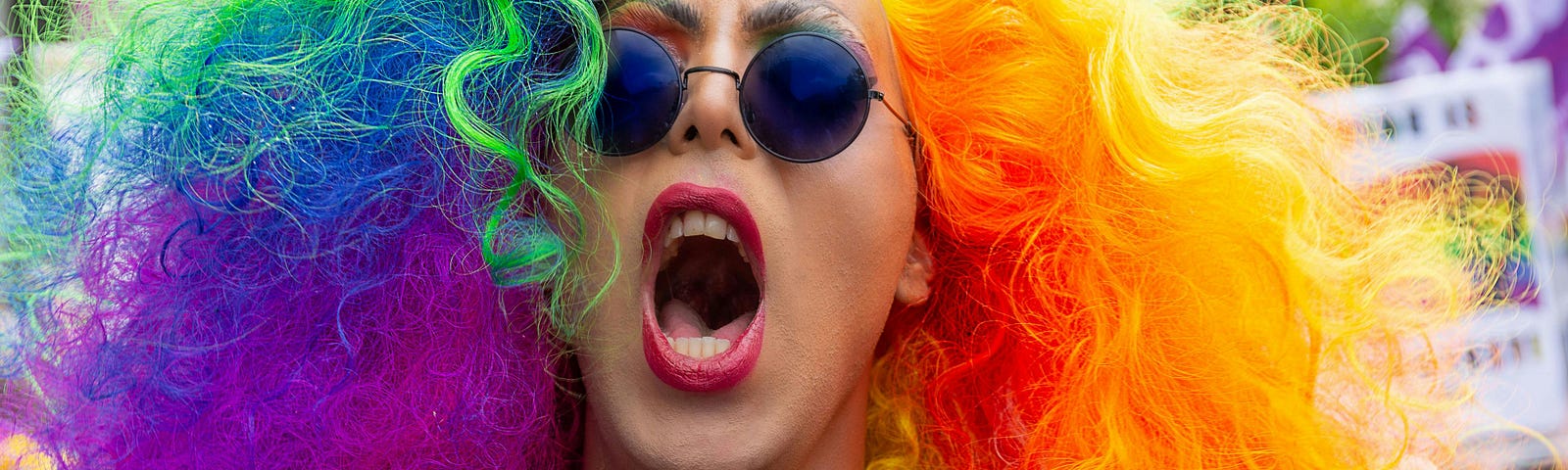 Headshot of person with long frizzy rainbow hair and eyeshadow, round sunglasses, and red lipstick yelling at the camera