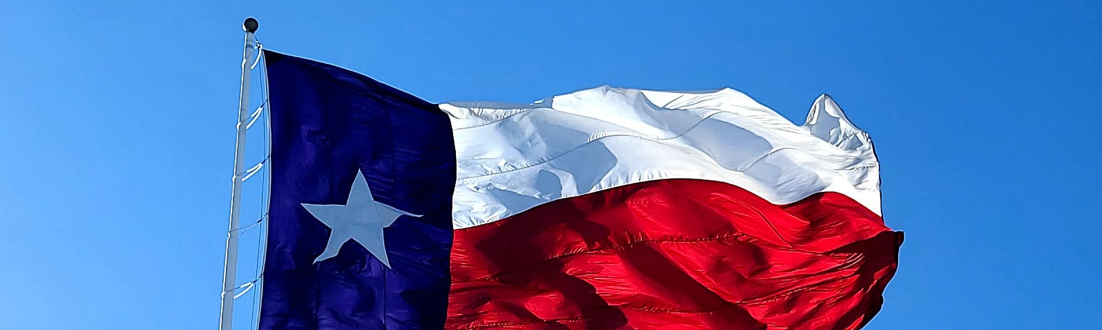 Texas Flag flying in the wind; one white star on a blue field with a large white section over a large red section.