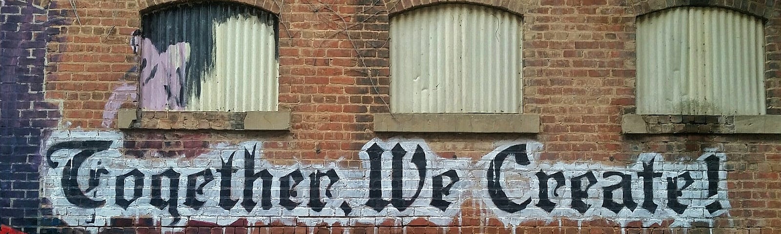 “Together, We Create!” spray painted in black lettering on an urban brick wall with shuttered windows.