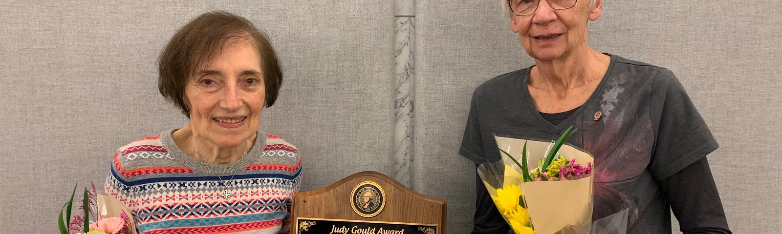 Two women smile, and hold flowers and a plaque that recognizes their awards.