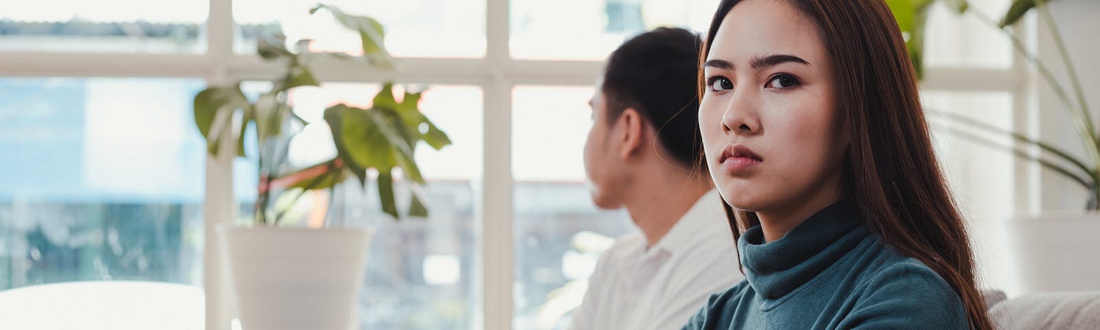 A young couple sits on a couch, their arms folded. One stares out of a window, while the other glares toward the camera.