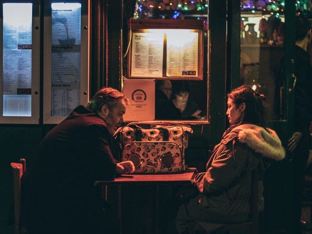 Photo Description: An older couple sit together in a dark diner having a discussion. They both look unhappy and still have their coats on. No food is on the table.