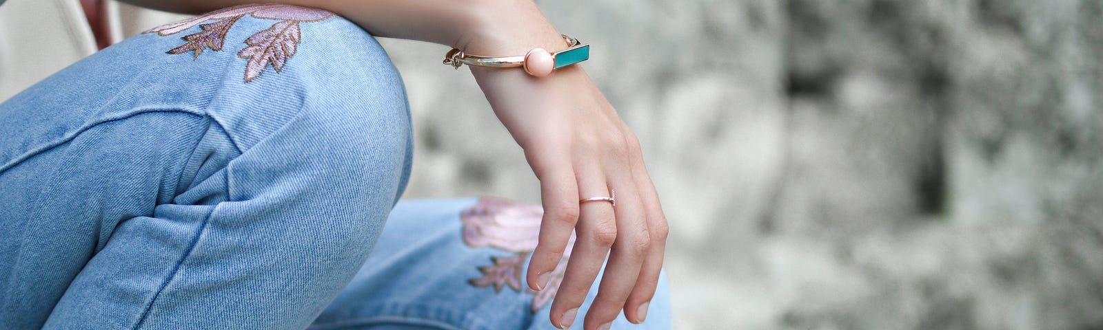 A young girl sitting down wearing jeans with pink flowers. She’s wearing a pink, gold and turquoise bracelet and a gold ring.